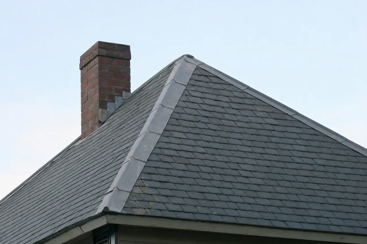 A hipped roof of grey-blue slates with lead-capped hips and a brick chimney against a pale sky — slate roofing, Sydney. Illustrative example.